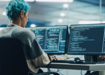 In Diverse Office: Young Stylish Woman in Working on Desktop Computer. Colorful Haired Girl Digital Entrepreneur Creating Modern Software, e-Commerce App Design, e-Business Coding. Over Shoulder Shot